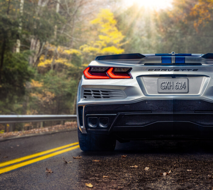 Close-up rear-view of 2024 Chevrolet Corvette E-Ray 3LZ convertible in Silver Flare with Electric Blue stripe package parked on a road in front of trees. Pre-production model shown. Actual production model may vary. Model year 2024 Corvette E-Ray available 2023.